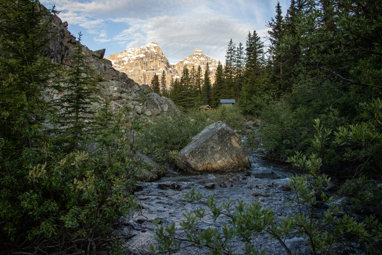 A stony braided mountain stream with a large boulder leads the eye to sunlit rocky summits above a dark spruce forest.