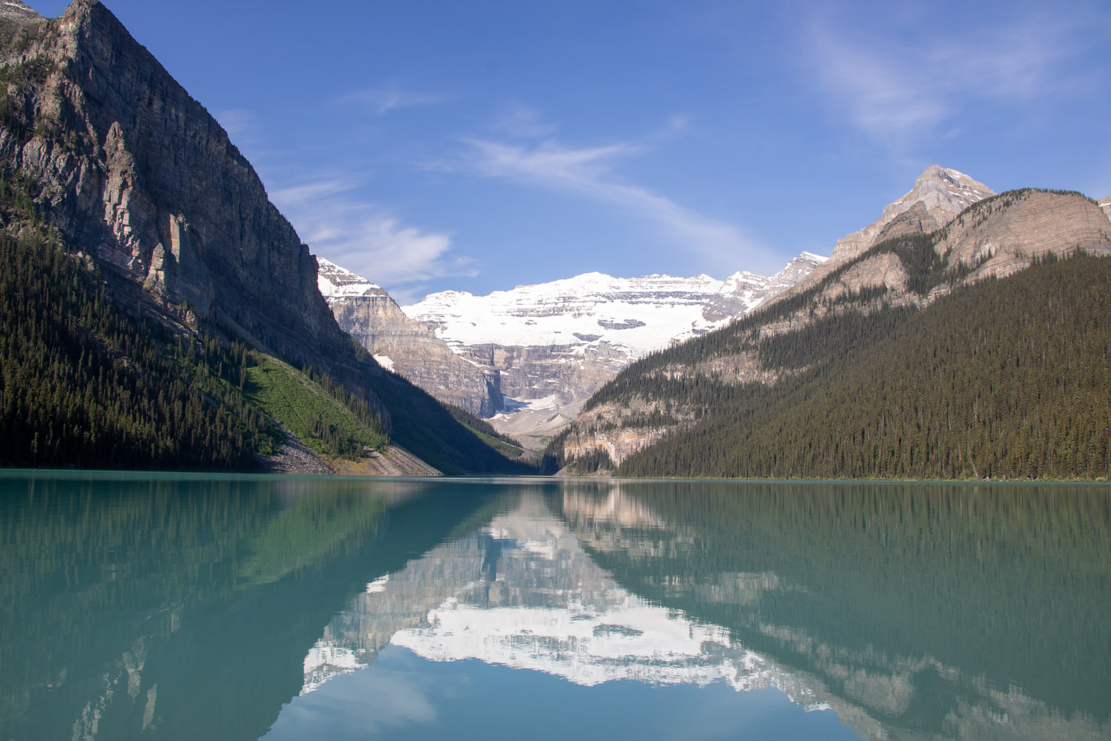 The snow-covered Victoria Glacier is reflected almost perfectly in the mirror-still turquoise water of Lake Louise between forested ridges.