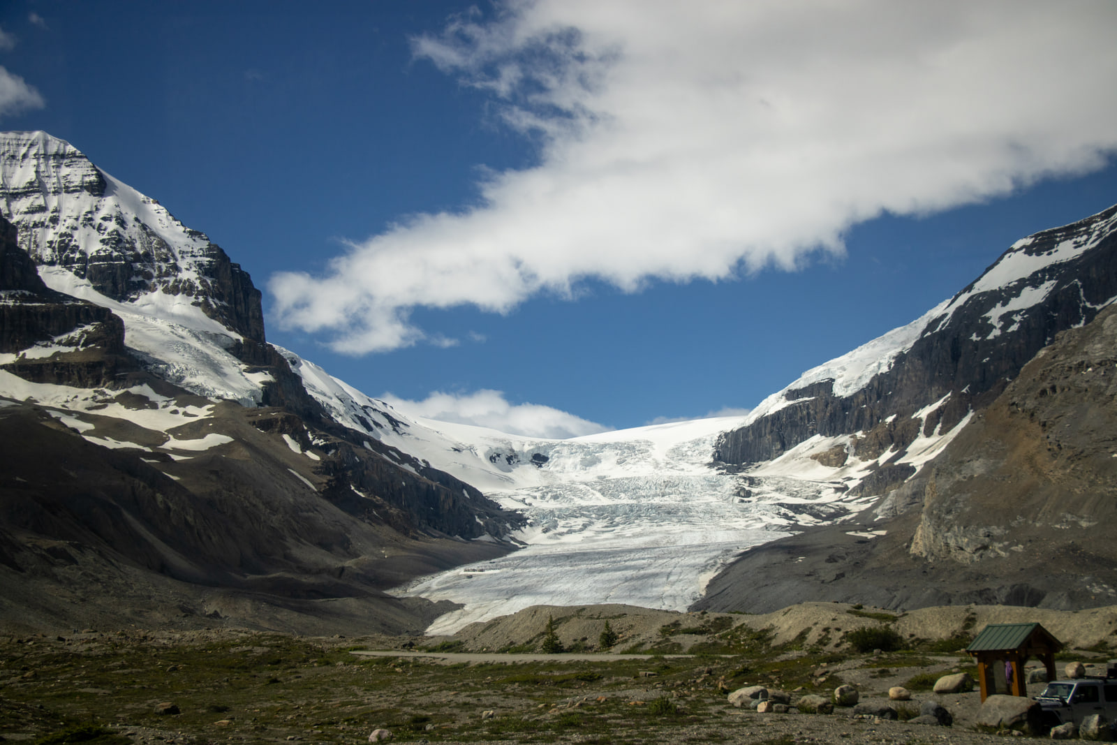 A glacier tongue descending between snow-streaked peaks under a band of cloud, with a small hut at lower right.