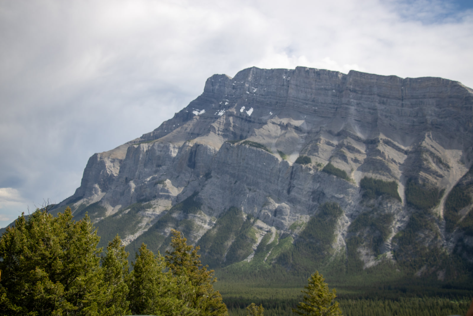 The diagonally layered rock face of Mount Rundle under heavy cloud, with a dark band of conifers along the base.