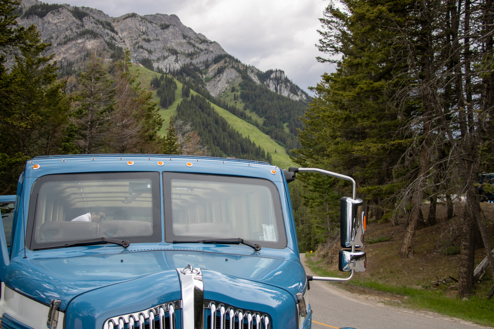 The chrome grille and windshield of a vintage light-blue truck parked below a grassy mountain slope and spruce forest.
