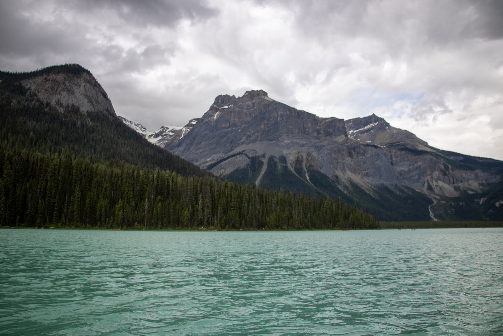 Milky green water of Emerald Lake in front of a dark spruce shoreline and a grey rocky peak under heavy cloud.