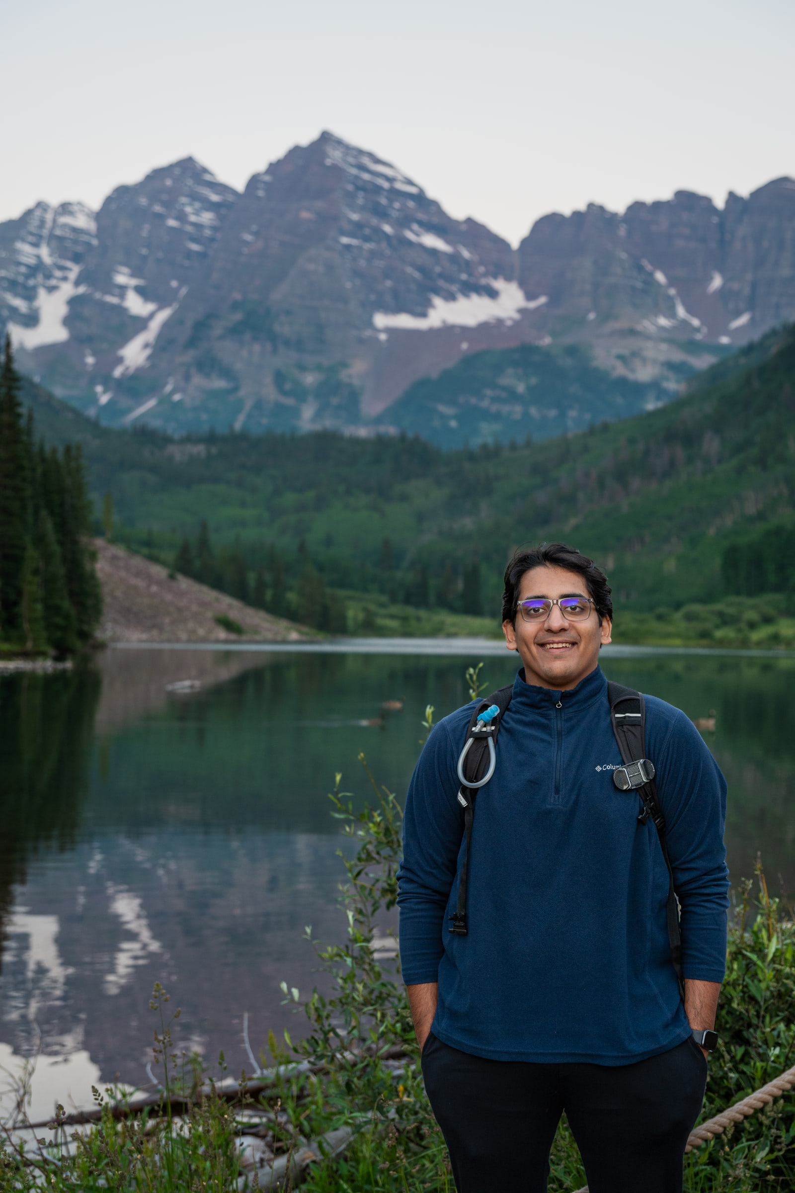 A man in a navy fleece and backpack stands smiling beside a still alpine lake with jagged peaks behind him at dawn.