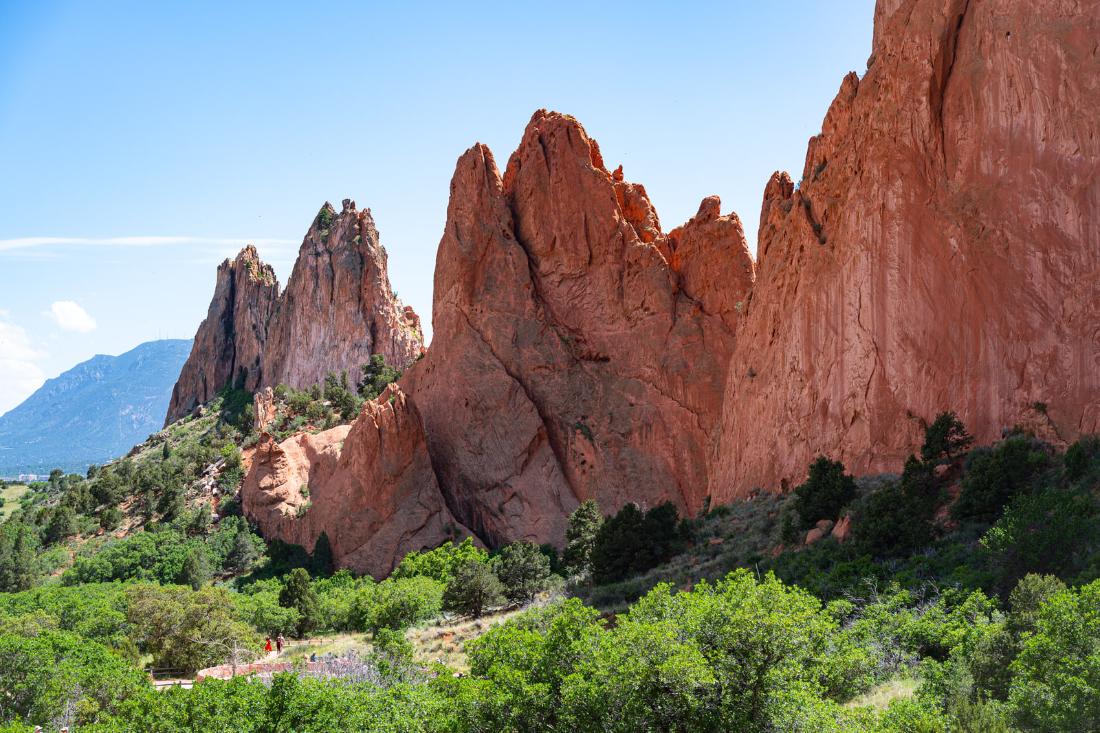 A huge fluted red sandstone fin towers over green scrub, with tiny hikers below and a distant blue mountain range behind.