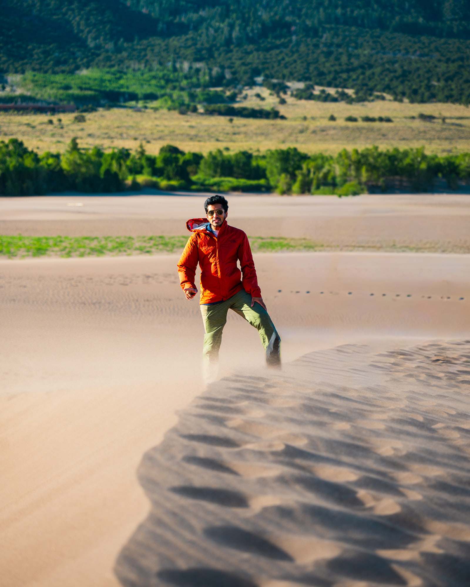 A man in an orange jacket drinks from a bottle while standing on a sunlit sand dune, with green hills and forest behind him.