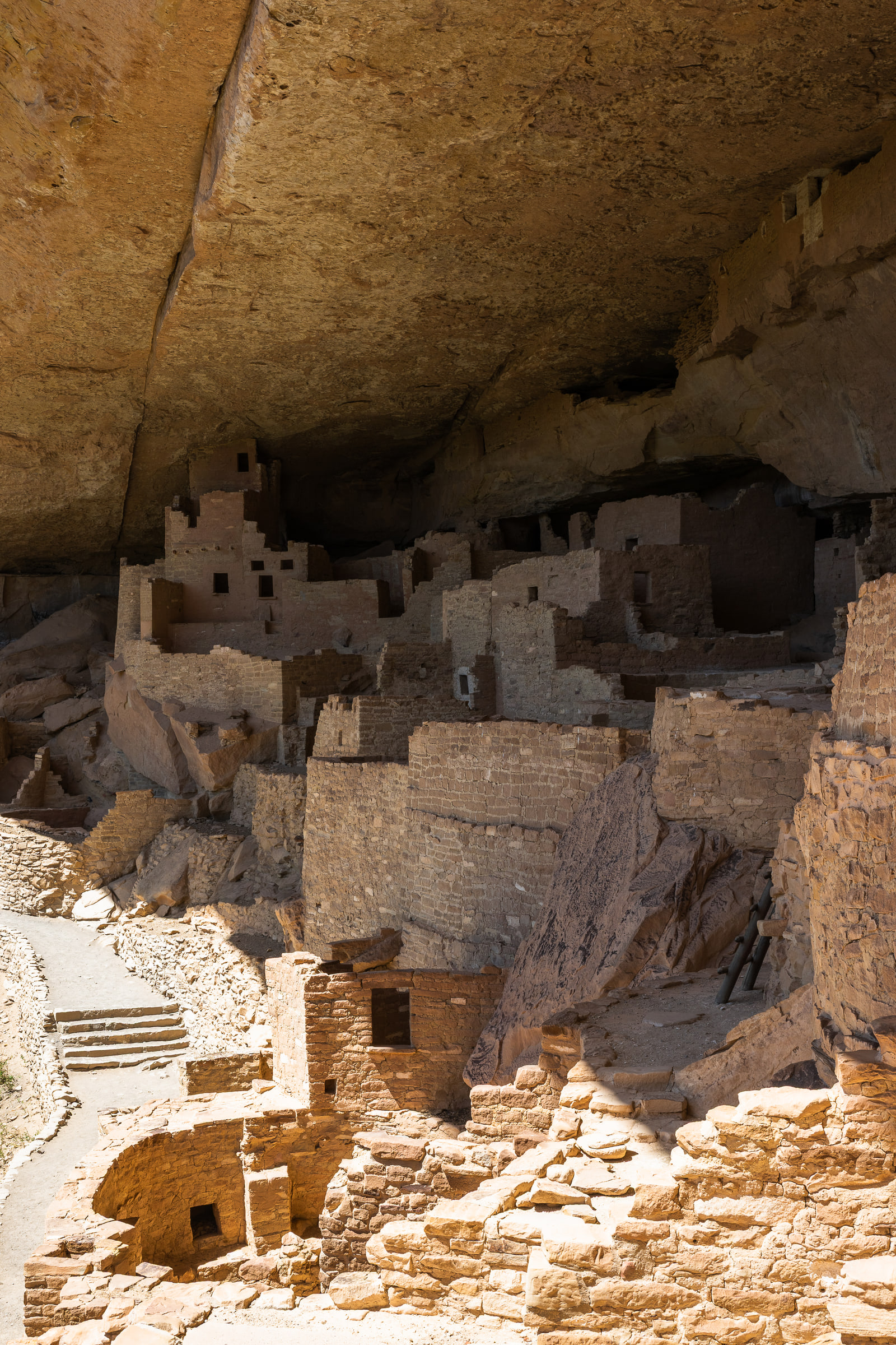 A vertical photograph of an ancient cliff dwelling sheltered beneath a vast curving sandstone overhang that fills the top of the frame.