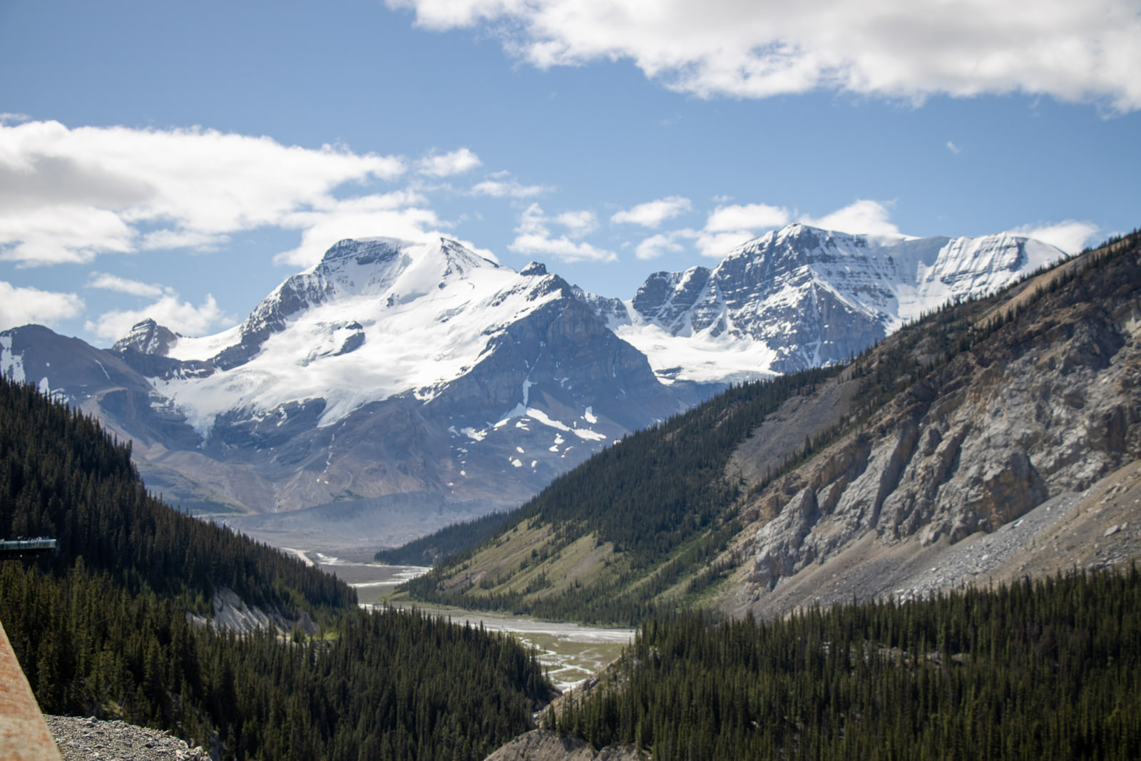 A wide glacial valley with a braided river leading toward a large snow-and-ice-covered mountain massif under scattered cumulus clouds.