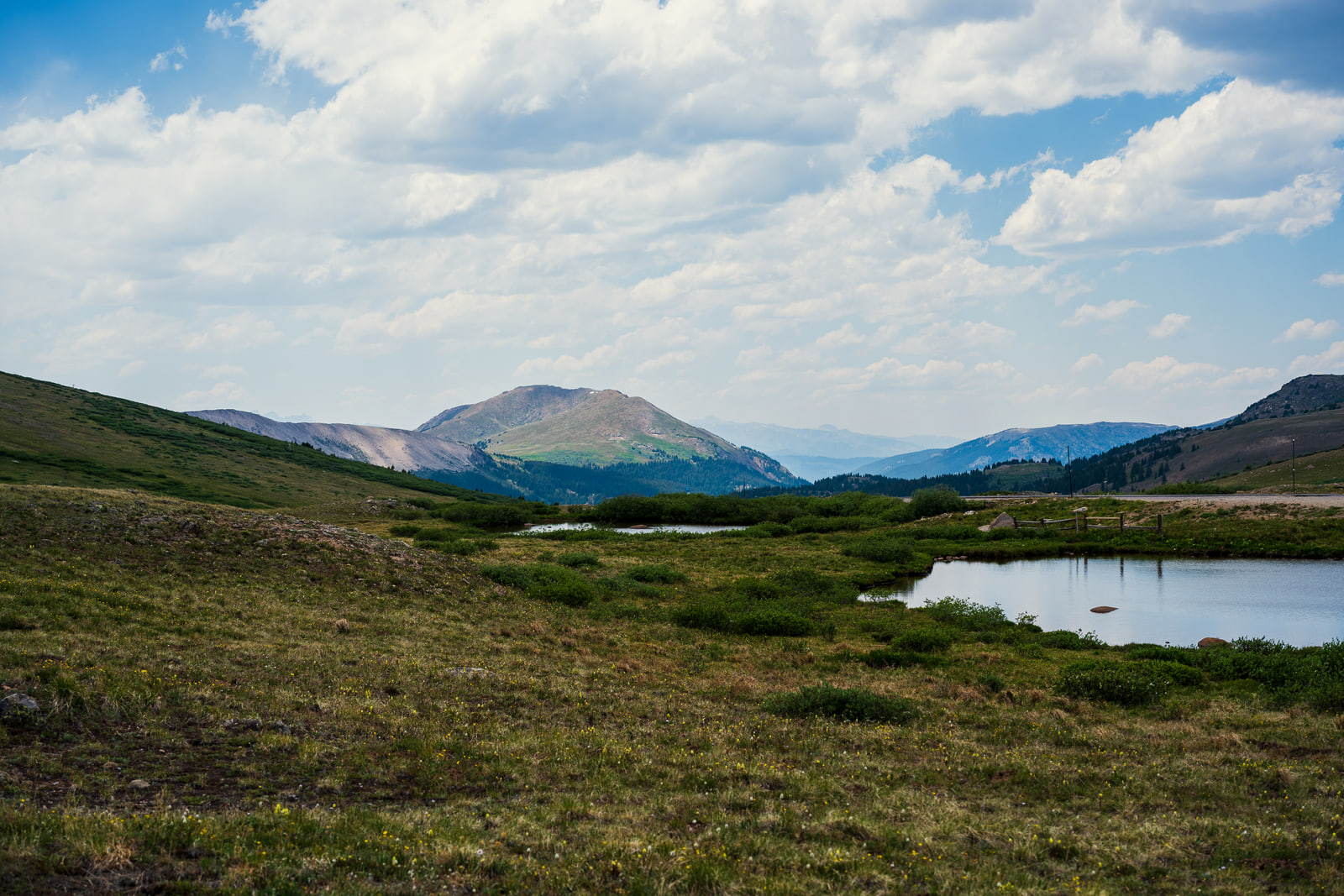 A small pond reflecting sky in an open alpine meadow, with a dark rounded peak and hazy blue mountains behind.