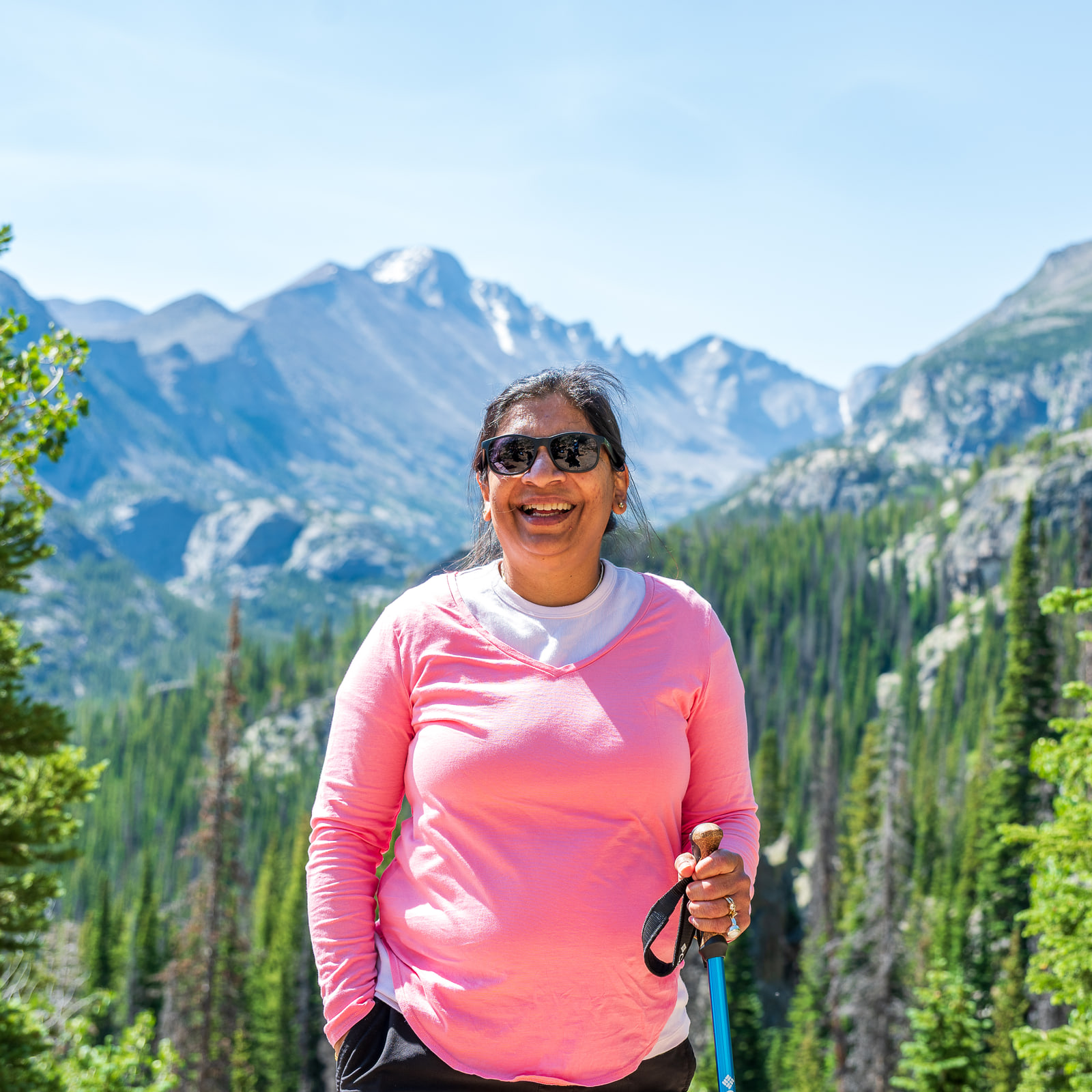 A smiling woman in a bright pink shirt and sunglasses holds a trekking pole in front of a snow-streaked granite mountain range.