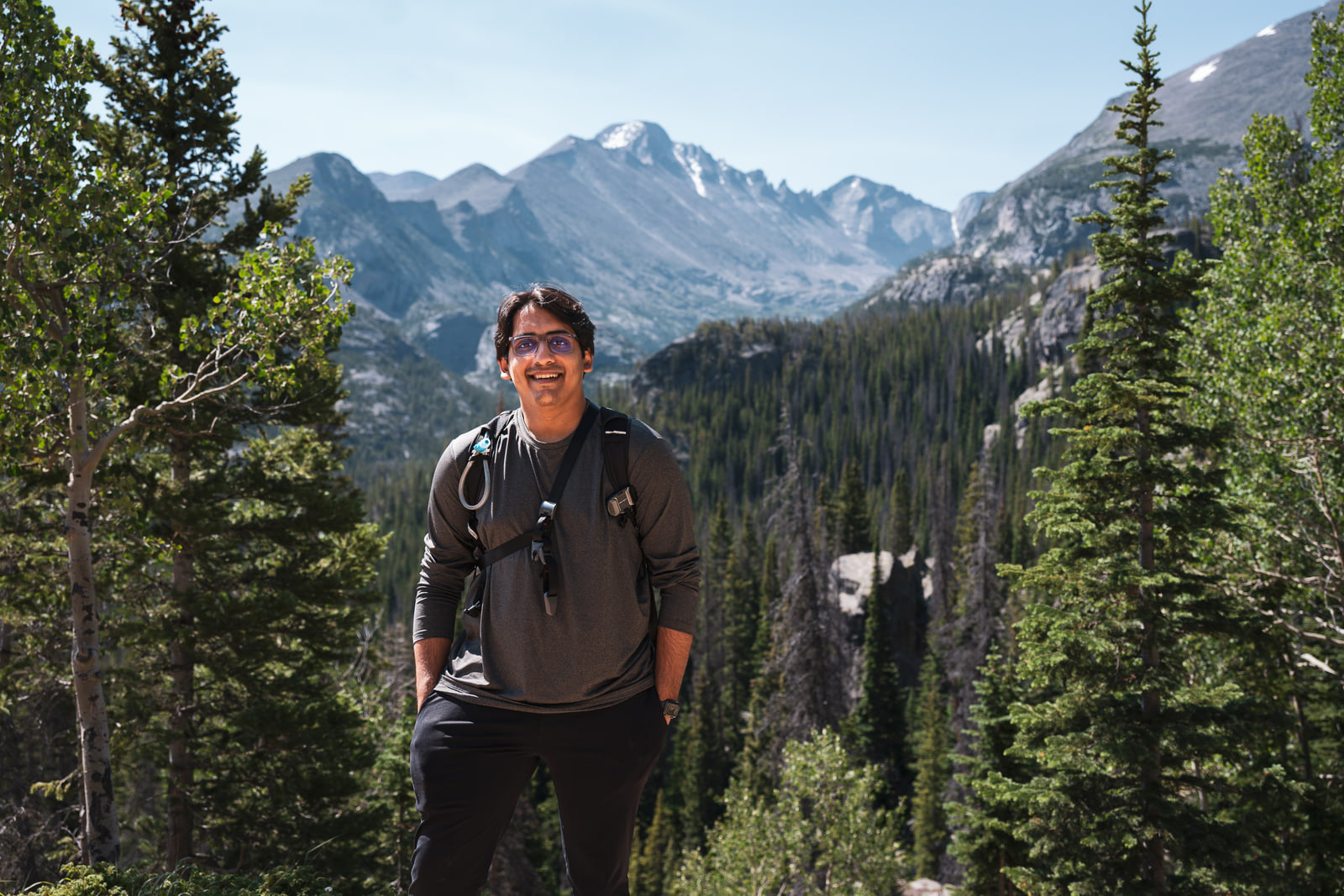 A young man with a daypack stands smiling between two spruce trees with a jagged snow-patched mountain range behind him.