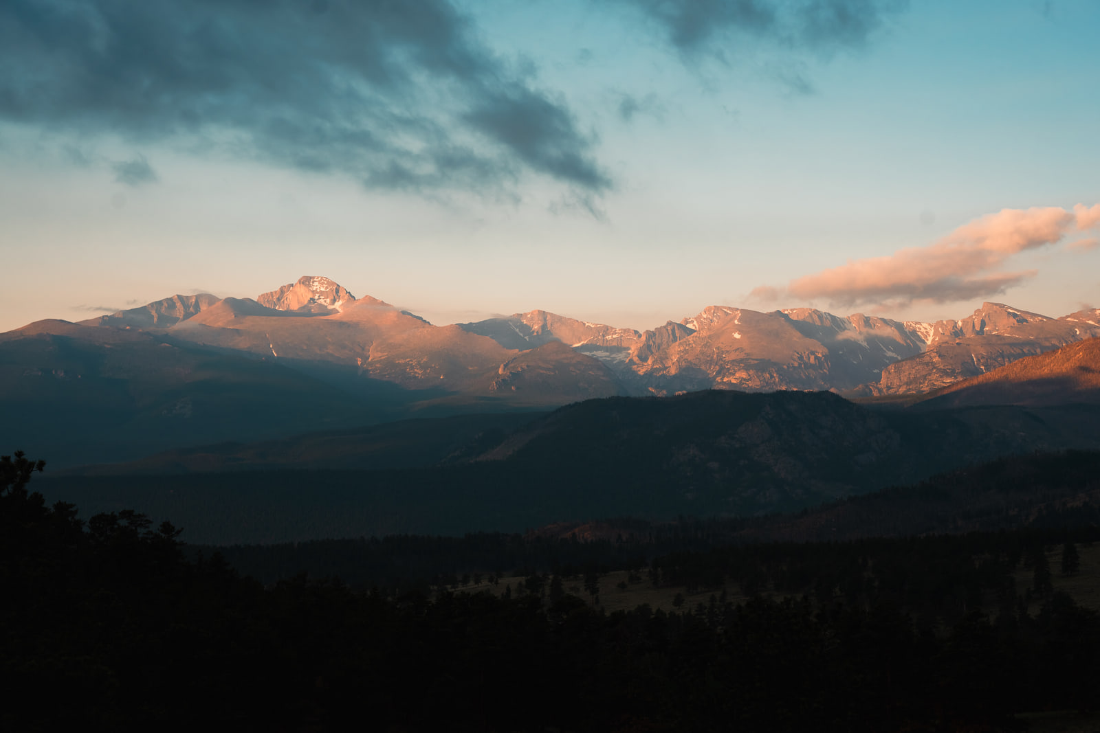 A sunlit mountain range at sunrise with a pink cloud streaming off the ridge above a dark forested valley.