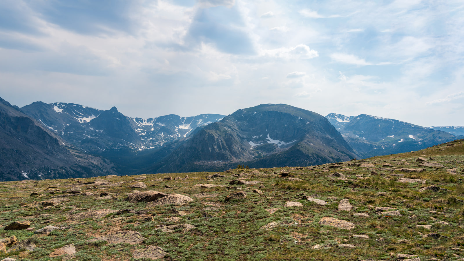 A panoramic view of rocky alpine tundra leading to snow-patched mountains under a bright cloudy sky.