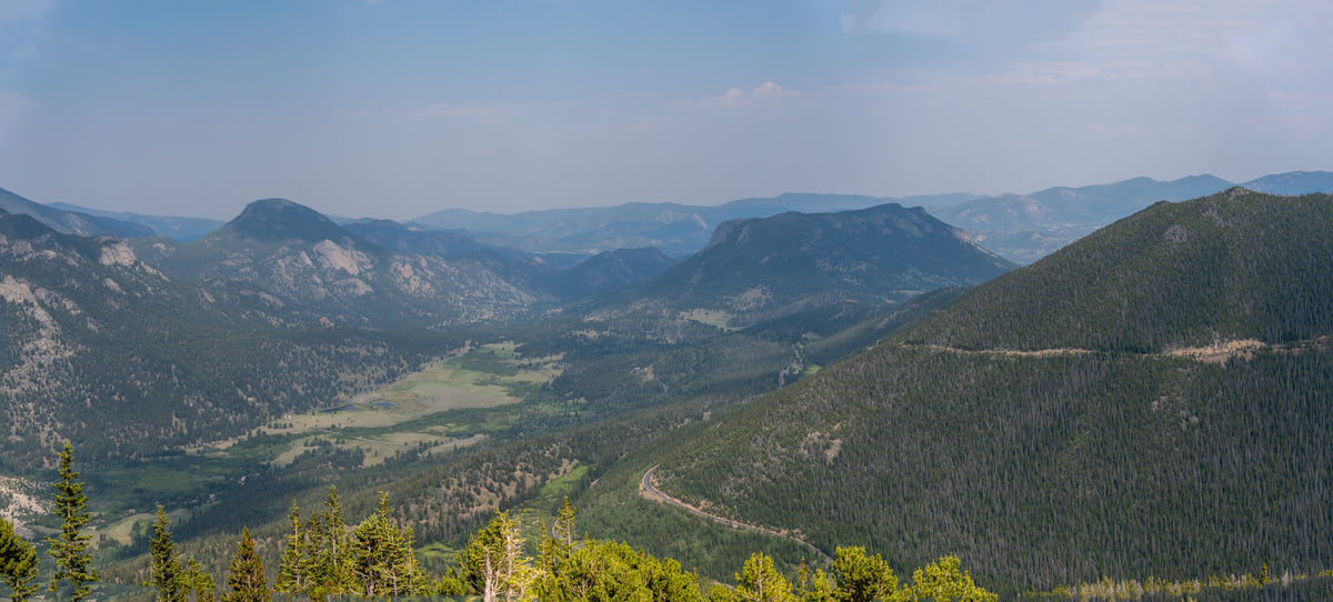 Panoramic view of a wide forested valley in the Colorado Rockies with layered mountain ridges fading into blue summer haze.