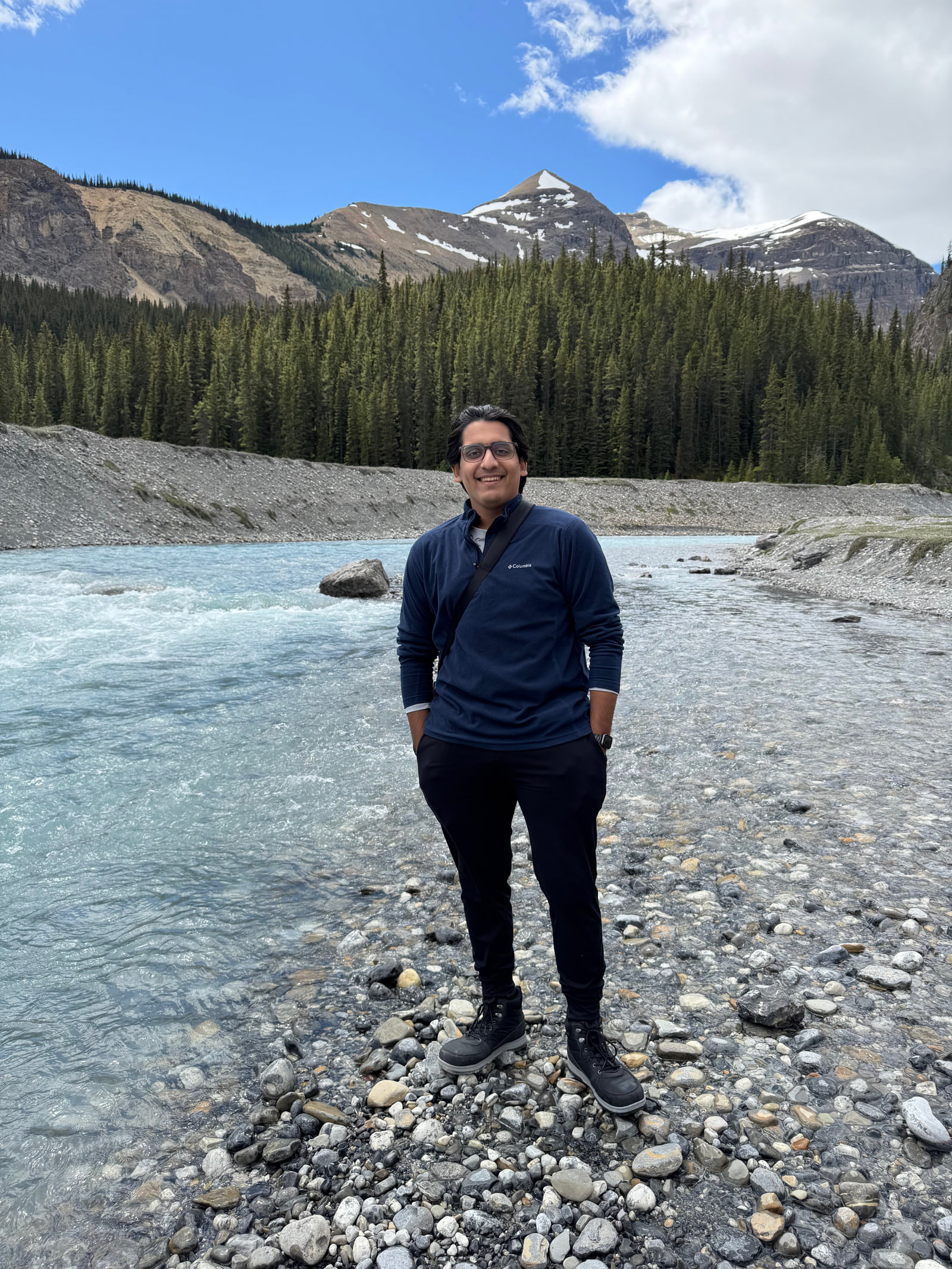 Aditya Shankar standing on the stony bank of a glacial river in the Canadian Rockies, spruce forest and a snow-streaked peak behind him.