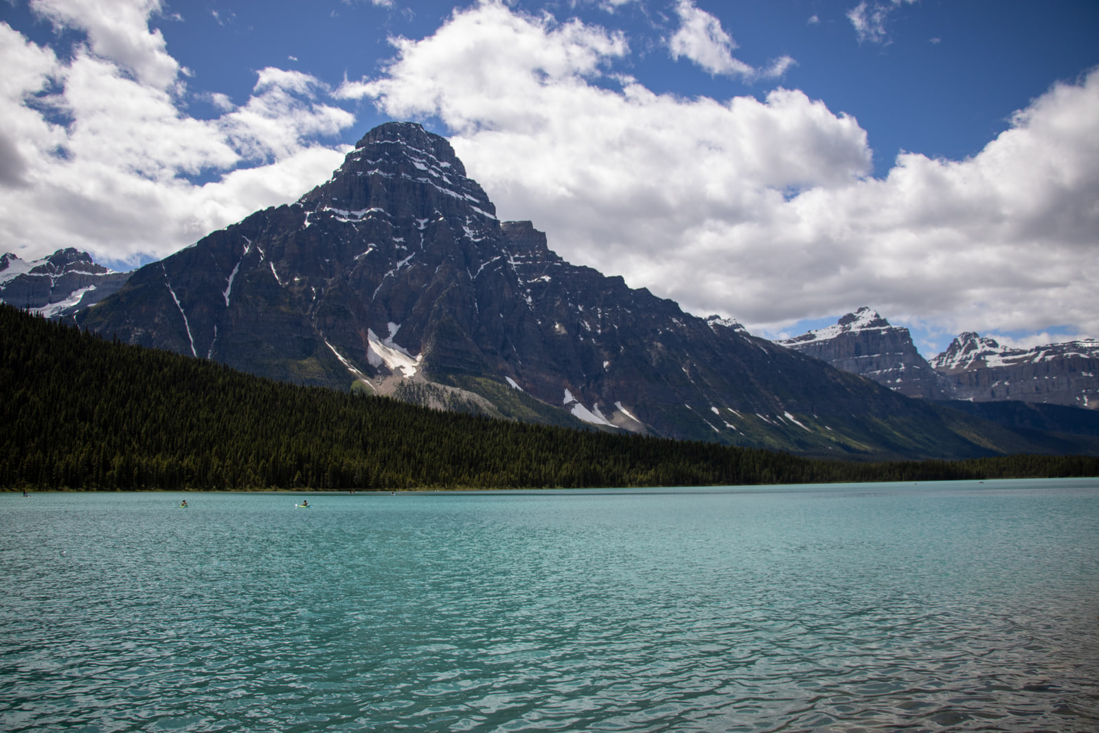 A steep dark pyramid-shaped mountain with a cloud-wrapped summit rising behind a turquoise glacial lake and dark spruce shoreline.