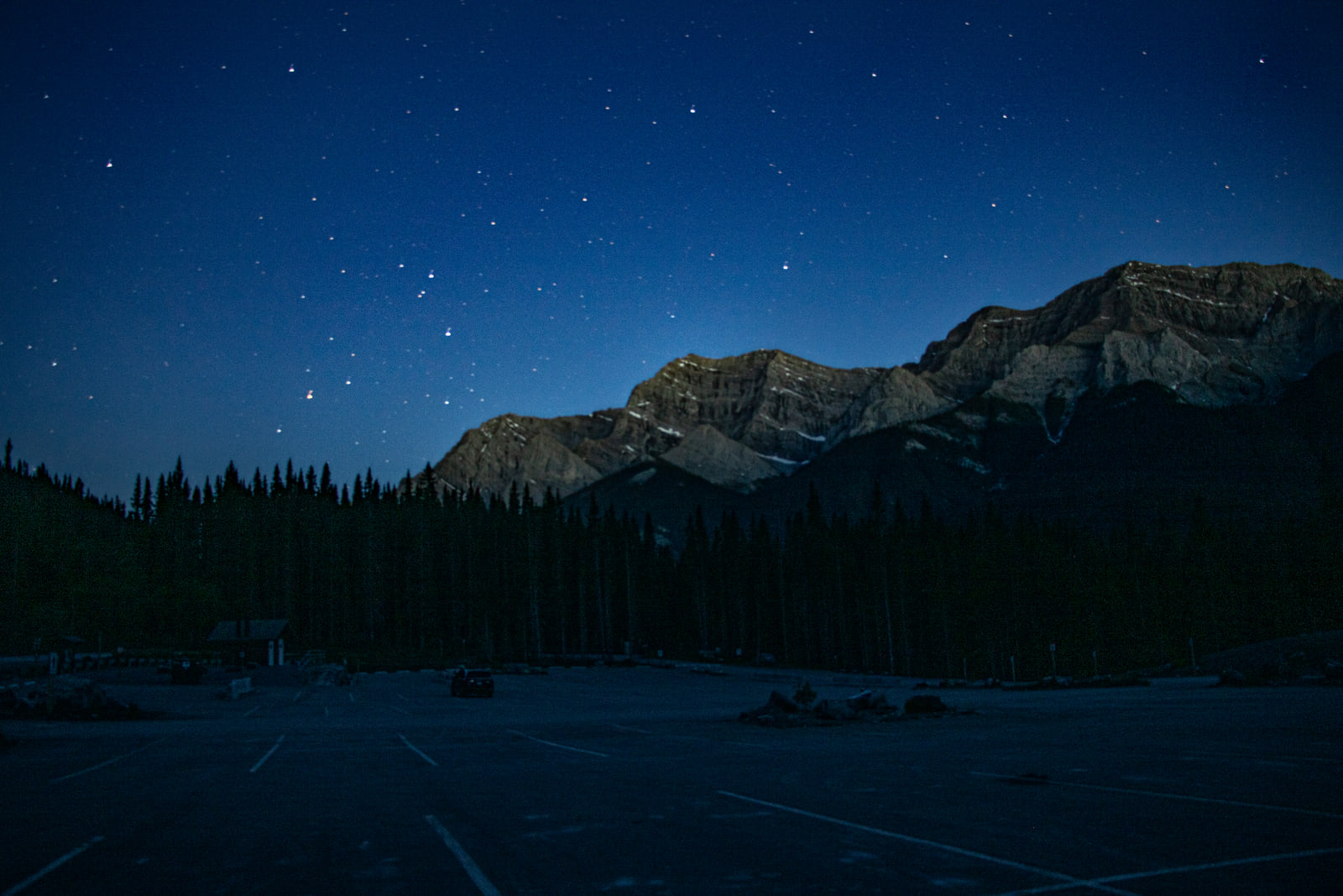 A star-filled night sky over a long layered mountain ridge, with a dark treeline and an empty parking lot in the foreground.