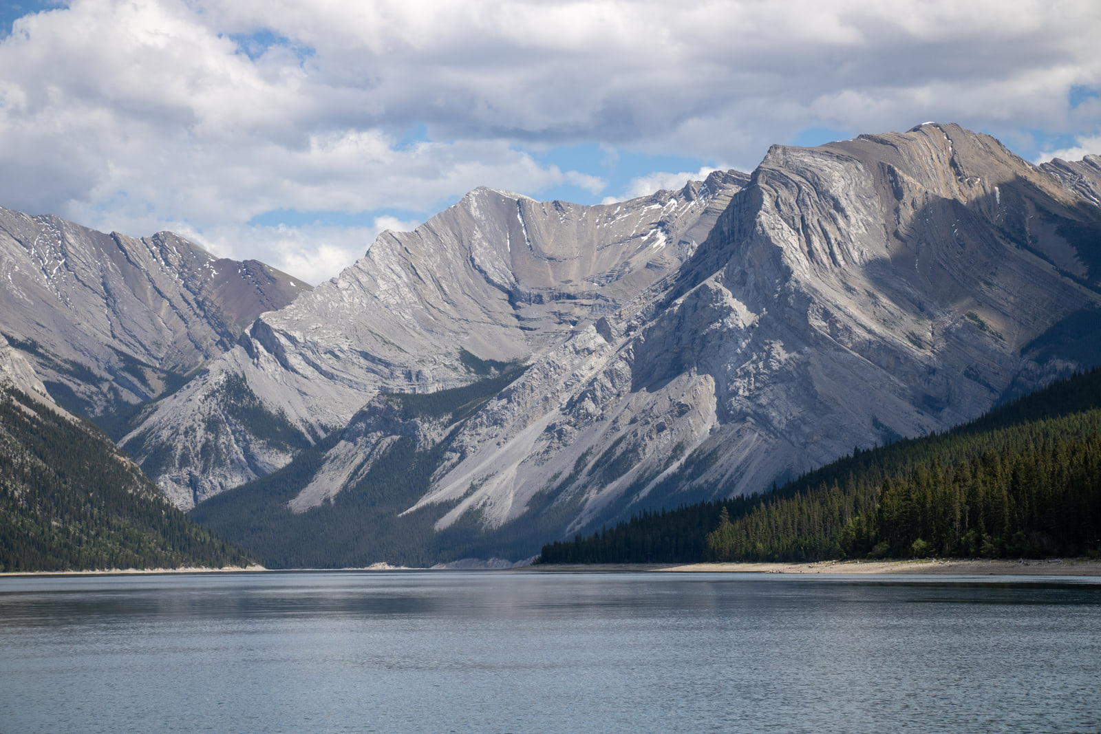 Steep grey folded mountain peaks rising above the dark calm water of a mountain lake, under large white cumulus clouds.