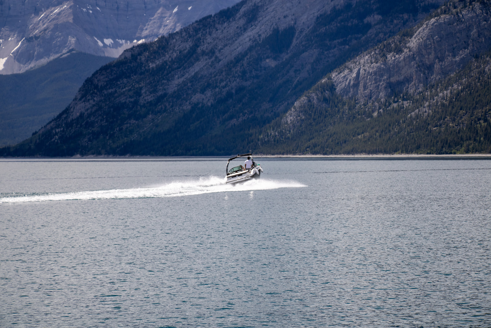 A speedboat crossing a wide pale lake and leaving a long white wake, with a dark mountainside rising behind it.