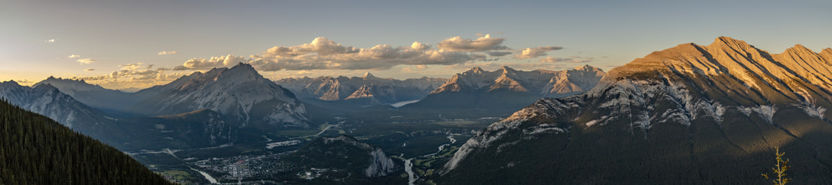 An ultra-wide panorama of the Bow Valley at golden hour, with the Banff townsite on the valley floor, a band of lit cloud over distant peaks, and a sunlit ridge at right.