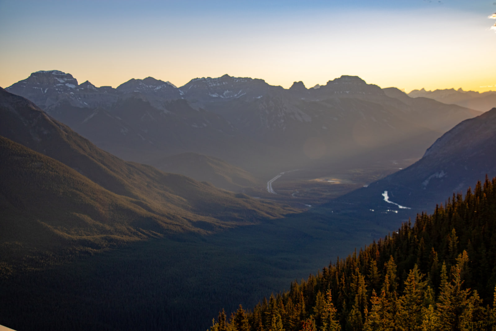 Layered mountain ridges stand in silhouette under an amber dusk sky while haze fills the valley below and a braided river catches the last light.