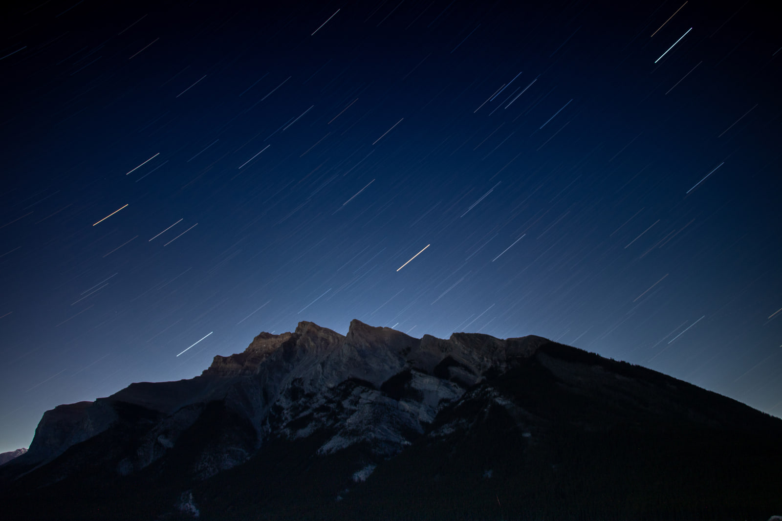 Short white star trails arc across a deep blue night sky above the dark, jagged silhouette of Mount Rundle in Banff.