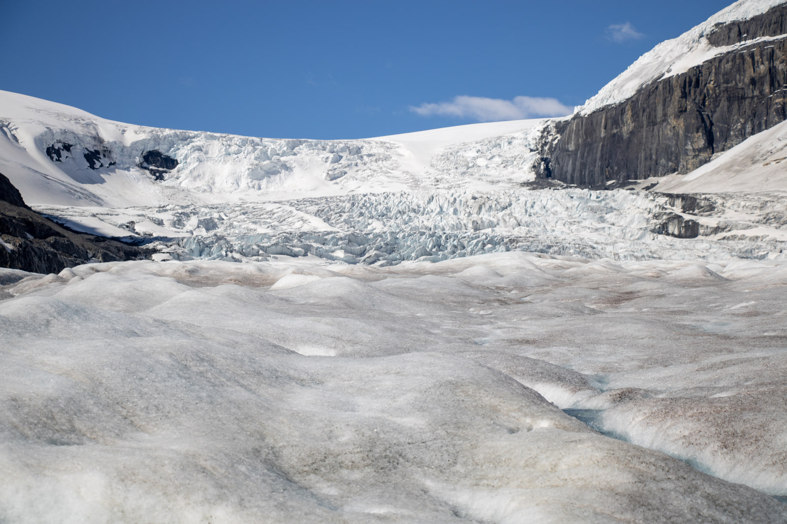 The rippled dirty surface of a glacier leading back to a blue crevassed icefall between rocky ridges.