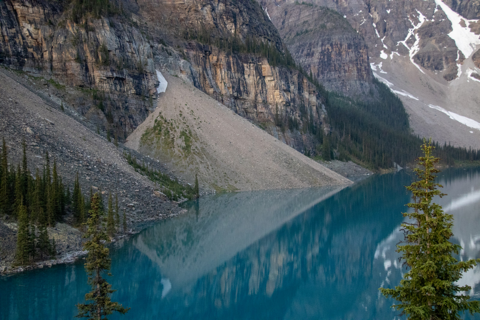 A pale scree slope and banded cliff dropping into bright turquoise lake water, with the slope's triangular shape repeating as a reflection.