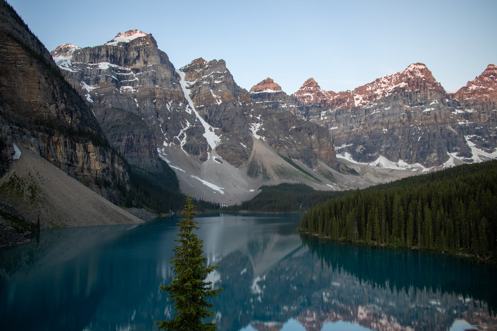 Sunrise alpenglow on the Valley of the Ten Peaks above Moraine Lake, the range mirrored in glassy turquoise water with a single spruce on the near shore.
