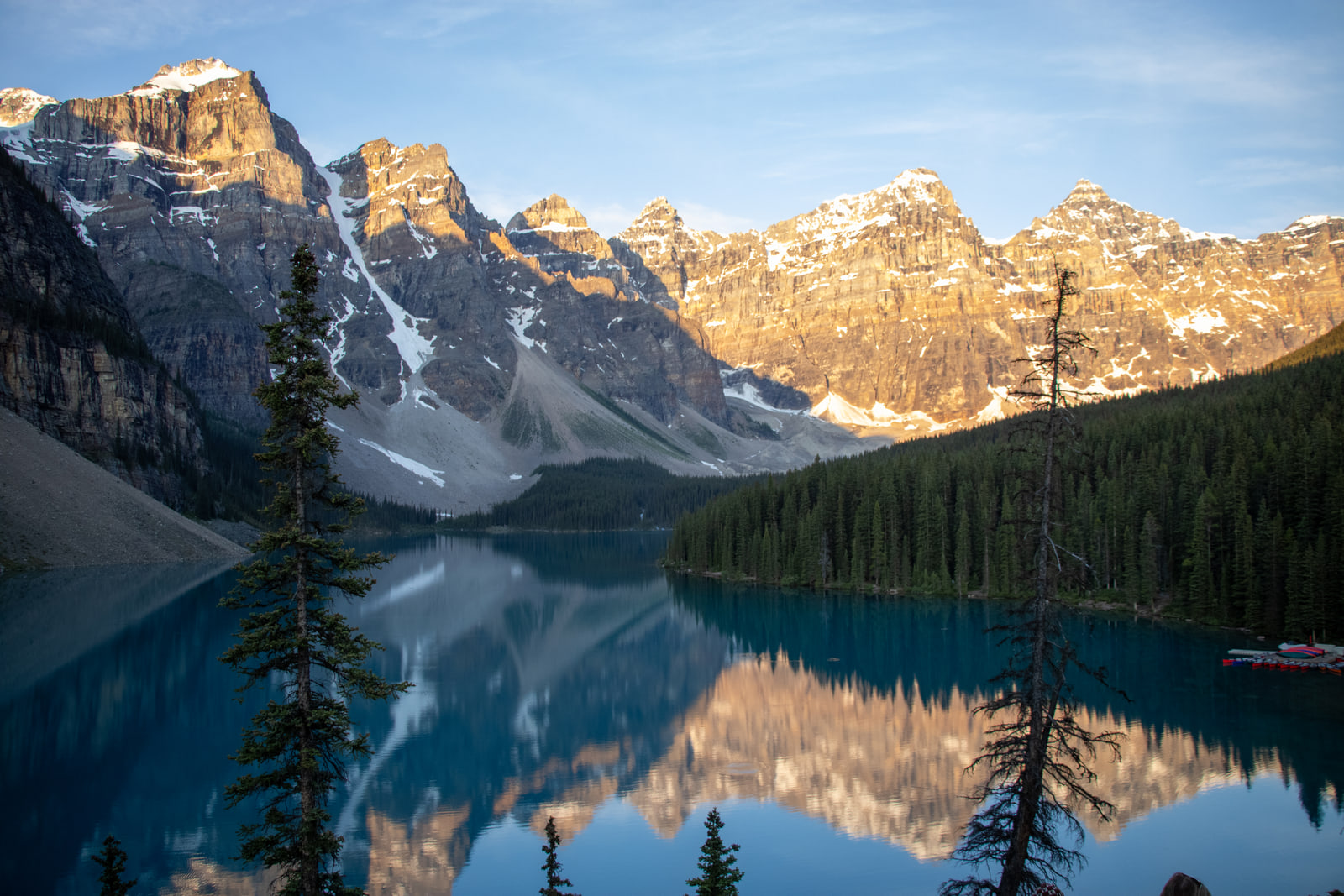 The Ten Peaks of Moraine Lake in full morning sun, reflected almost perfectly in glassy turquoise water and framed by dark spruce silhouettes.