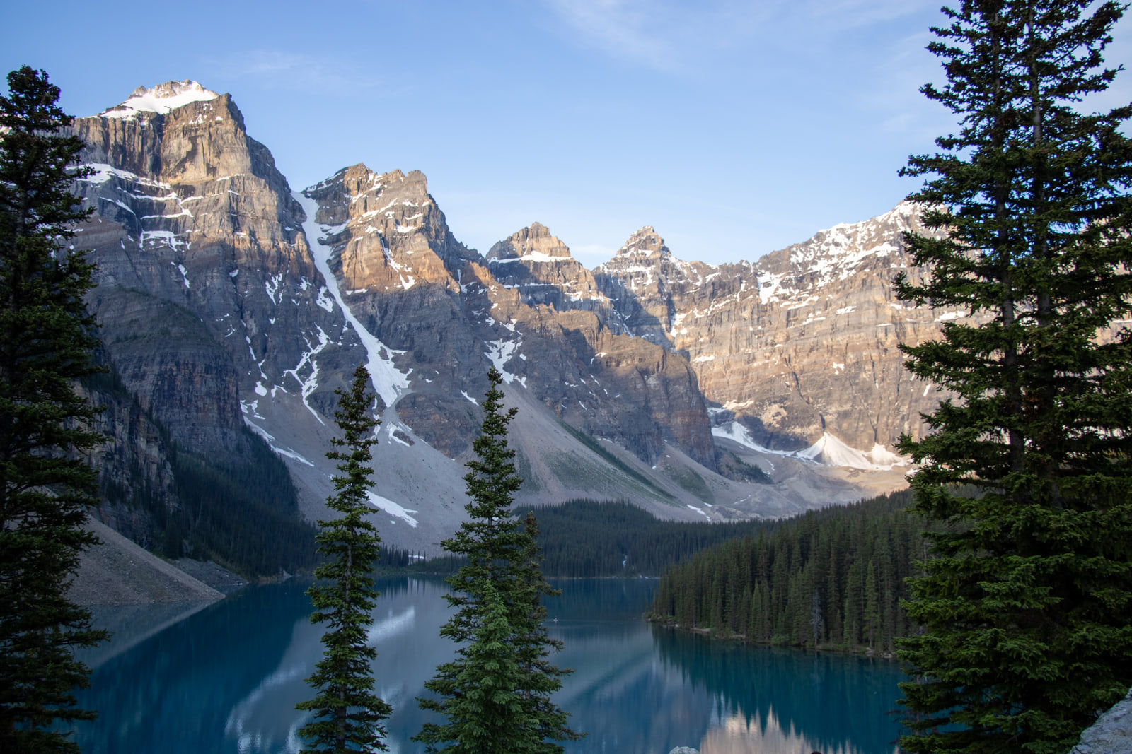 Sunlit jagged peaks of the Valley of the Ten Peaks rise above the shadowed turquoise water of Moraine Lake, framed by dark spruce trees.